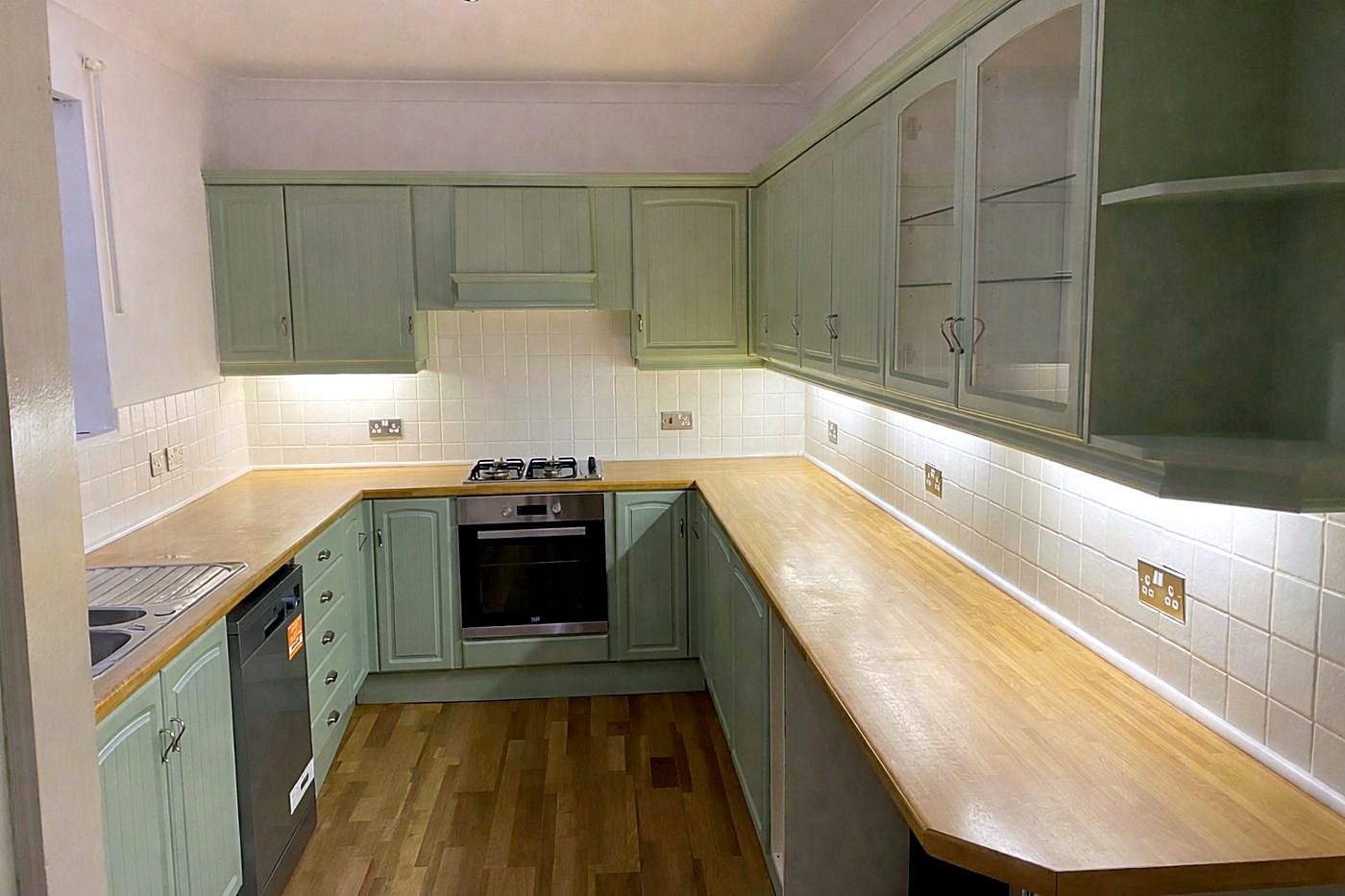 The same kitchen after: sage green units, white tiling, wood-effect floor and under-cabinet lights